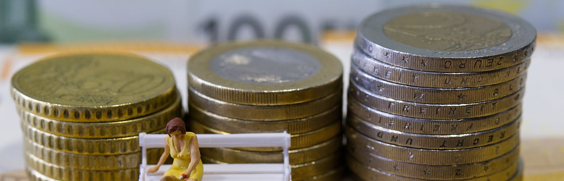 Eine miniatur Bank mit Mutter und Kind. Im Hintergrund gestapelte Geldmünzen.