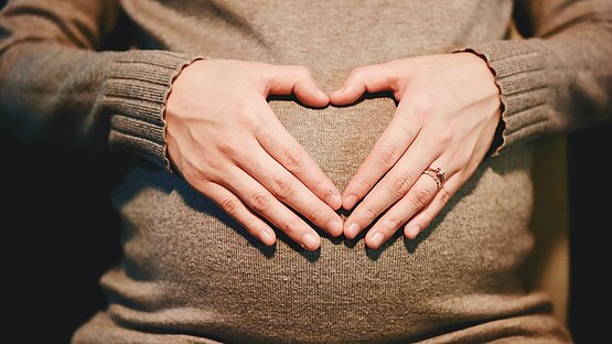 Schwangere Frau hält Hände vor ihren Bauch. Hände bilden eine Herzform.